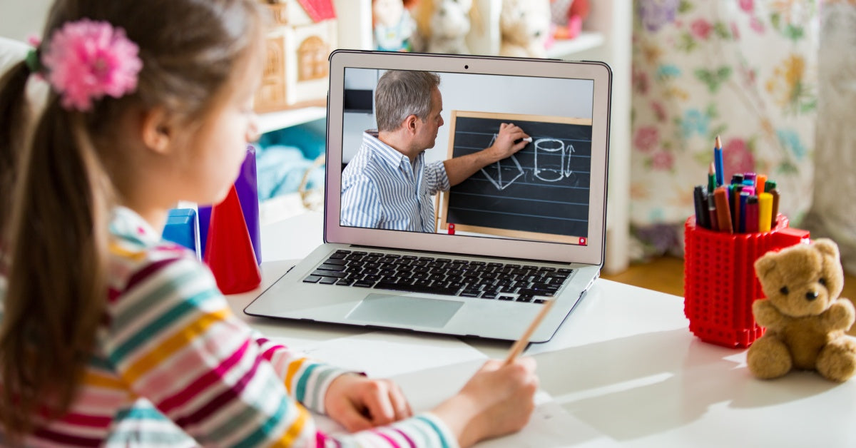 A young girl attends a remote learning session inside her bedroom. She watches her teacher draw geometric shapes on a chalkboard.