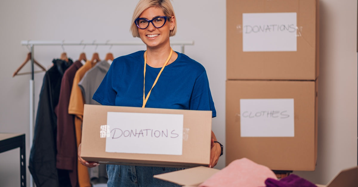 A smiling woman in glasses and a blue shirt holds a box labeled donations. There are more boxes in the background.