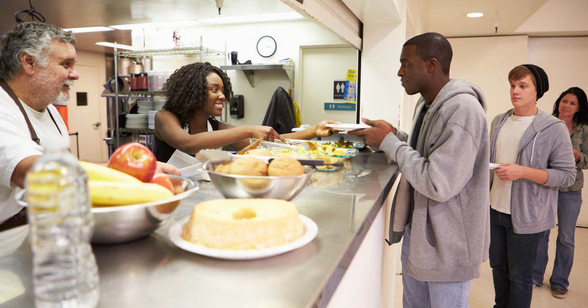 A smiling black woman and a white man handing out food to the people lined up. There is fruit, a bottle of water, and bread.