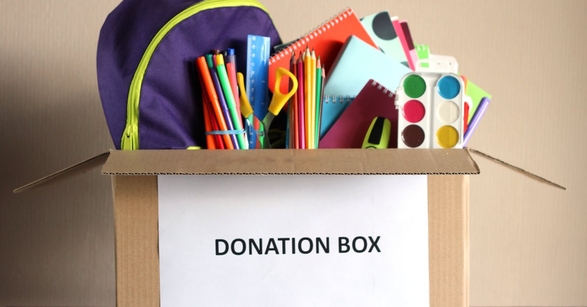 A cardboard box with a piece of paper with the words 'donation box' typed on it, full of a backpack and school supplies.