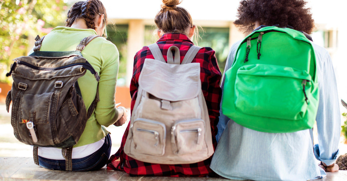 Three young people with their backs to the camera are wearing green and gray backpacks while sitting on a wall outside.