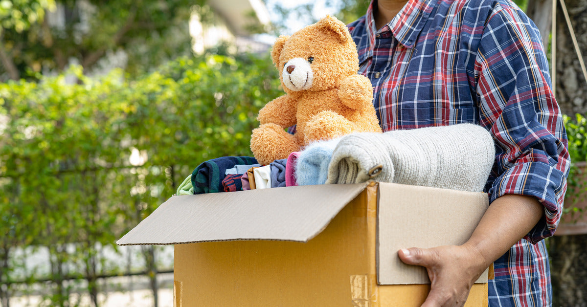 A person is carrying a small box of folded clothes and sweaters. A teddy bear sits on top of the items in the box.