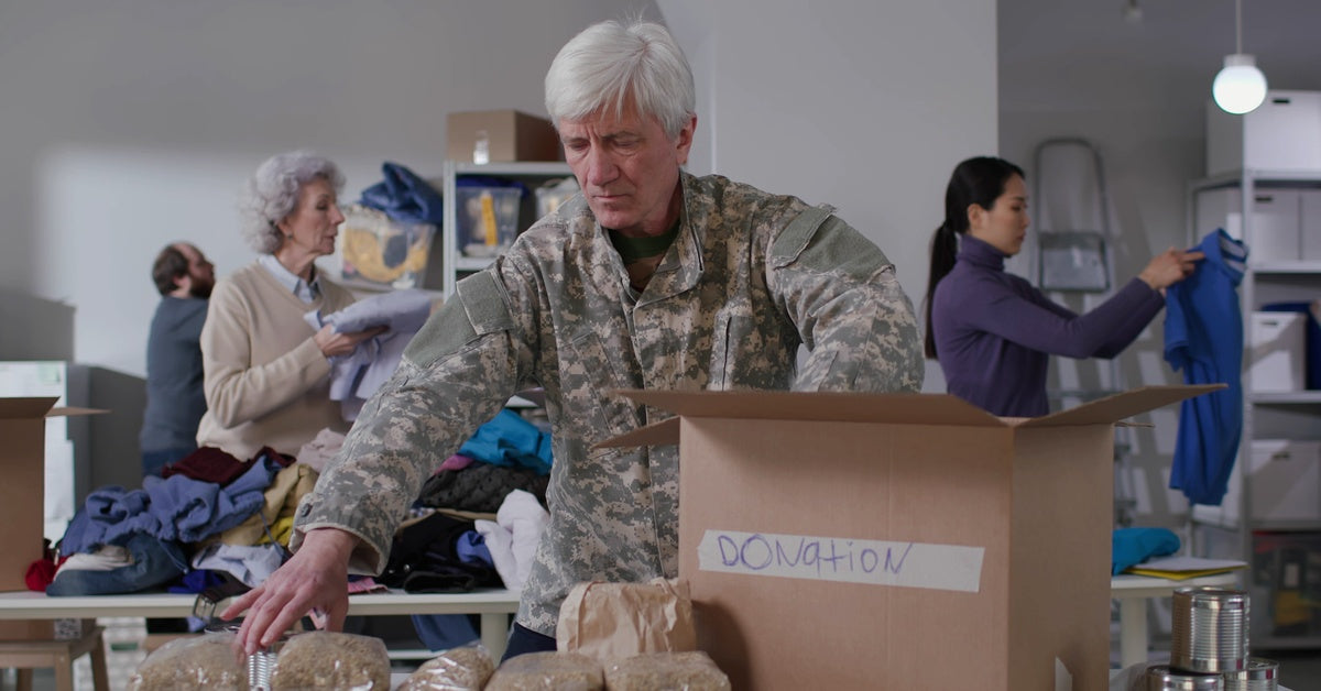 A man in military uniform packs supplies into a box labeled "donation." People fold clothes in the background.